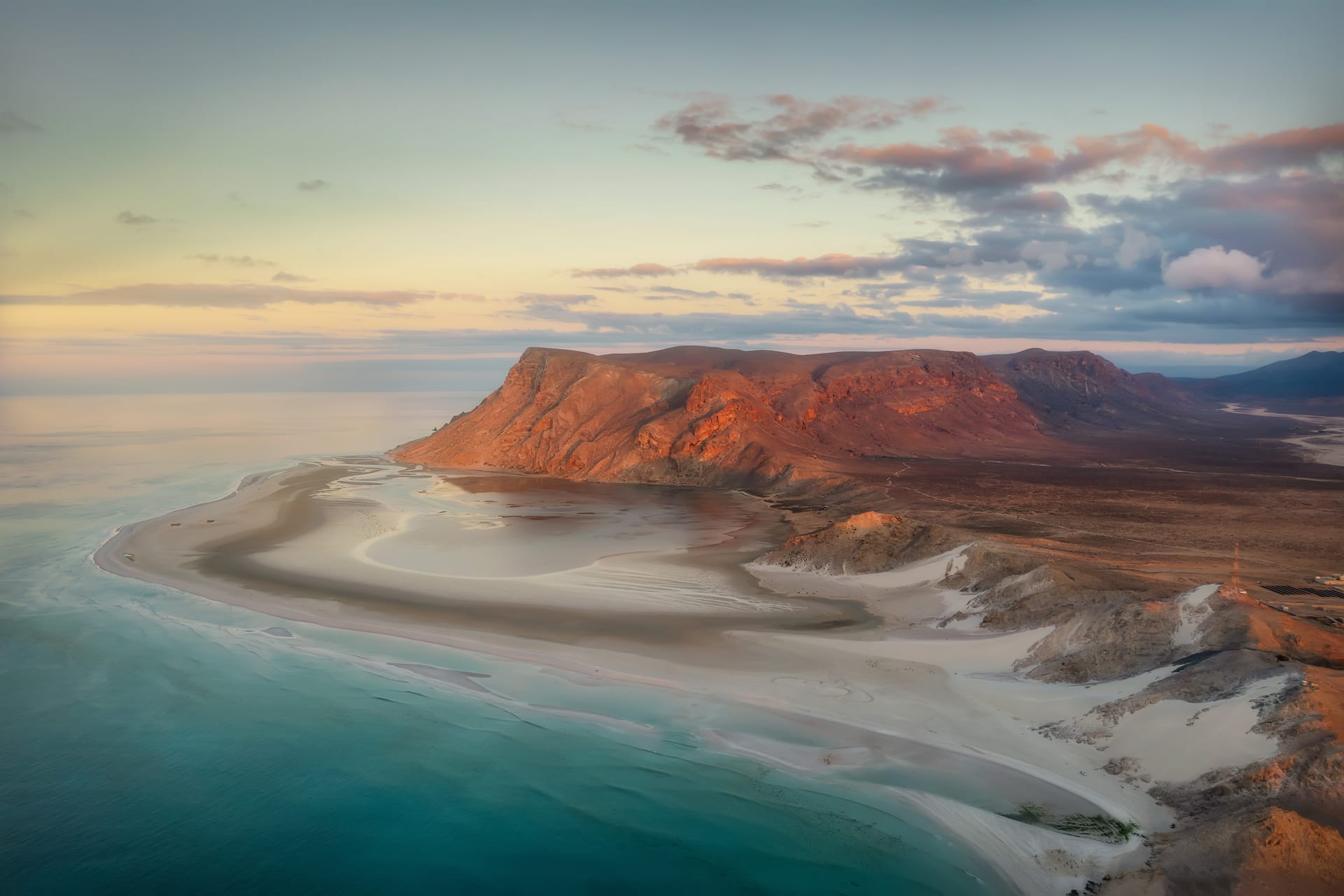 Detwah Lagoon from above at dusk, pale sand bars curving through shallow water below a red headland.