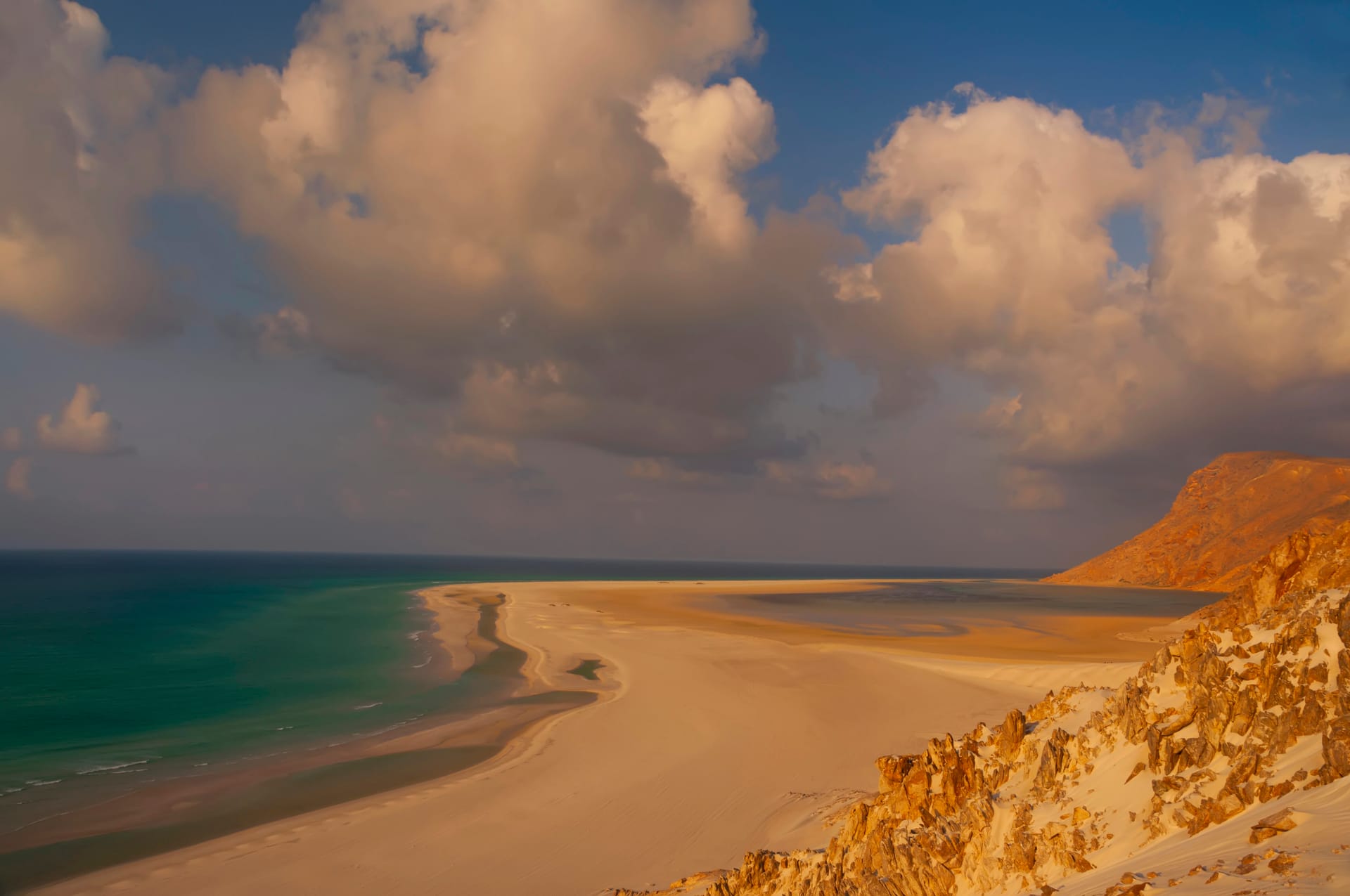 Sandy slopes falling to a curved beach and a calm sea under large clouds.